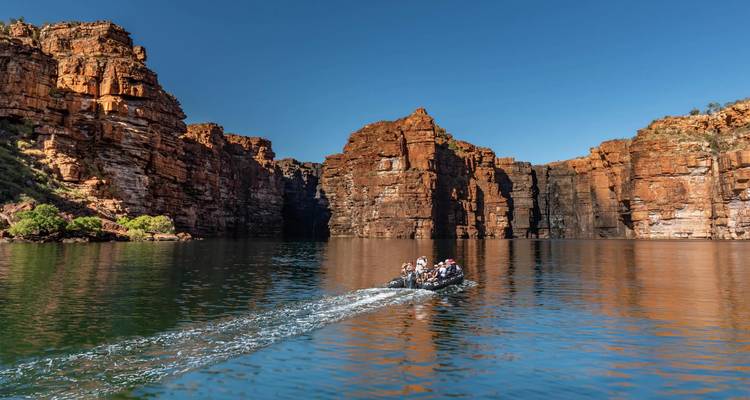 Zodiac met reizigers die door spiegelglad water varen onder de steile roestkleurige canyonwanden van Kimberley.