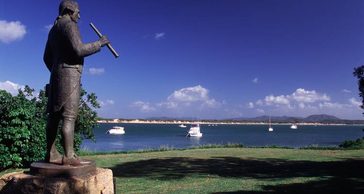 Bronzestatue von Captain Cook blickt über eine ruhige Bucht mit vor Anker liegenden Booten und fernen Hügeln.