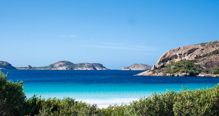 El mar turquesa brillante se encuentra con la playa de arena blanca y los islotes de granito distantes bajo un cielo sin nubes.