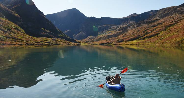 A lone paddler in an inflatable kayak glides across a crystal alpine lake encircled by steep green and rocky mountains.