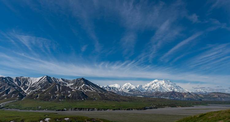 A panoramic view of Denali’s snow-covered peaks rising above green tundra under dramatic streaking clouds.