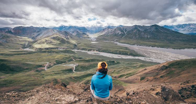 A hiker in a blue jacket sits on a rocky ridge overlooking a vast valley of winding rivers and mountains beneath dramatic clouds.