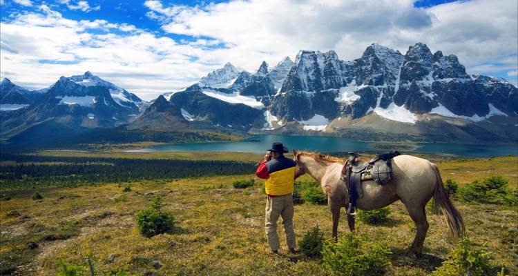 A lone rider in colourful outdoor gear stands beside a saddled horse, gazing at a turquoise lake and jagged snowy peaks.