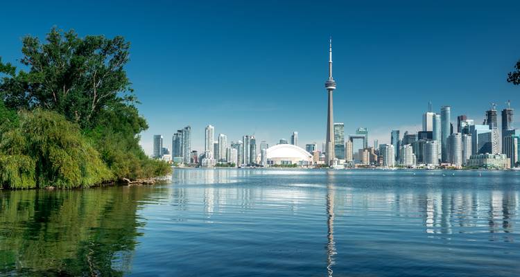 El horizonte de Toronto con la Torre CN y el Rogers Centre reflejados en las aguas tranquilas del Lago Ontario bajo un cielo azul despejado.