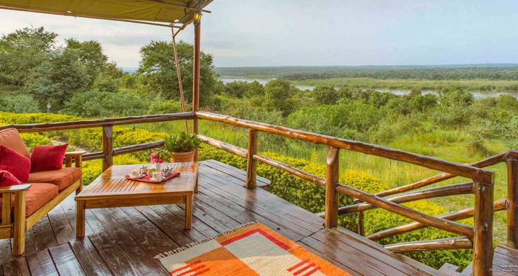 Terrasse en bois avec coussins colorés donnant sur une zone humide luxuriante et un lac au loin dans un cadre de lodge de safari.