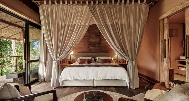 Suite de lodge safari élégante avec un lit à baldaquin, des accents en bois naturel et une vue sur la forêt à travers de grandes fenêtres.