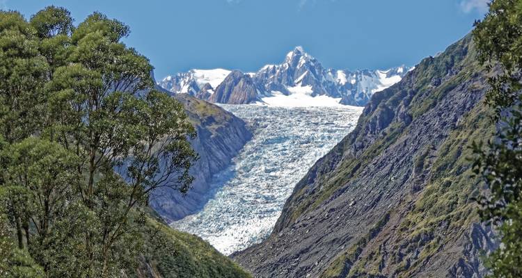 Panoramablick auf einen Gletscher, der zwischen schroffen Gipfeln hinabfließt, mit schneebedeckten Bergen in der Ferne.