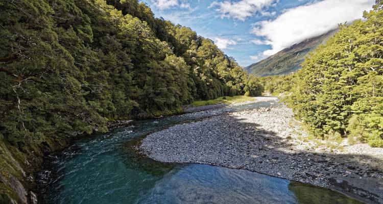 Klarer smaragdgrüner Fluss, der sich durch ein dicht bewaldetes Tal unter einem strahlend blauen Himmel mit leichten Wolken schlängelt.