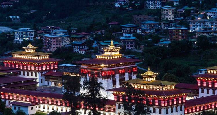 Nachtansicht des beleuchteten bhutanischen Dzong-Komplexes mit Stadtlichtern ringsum.