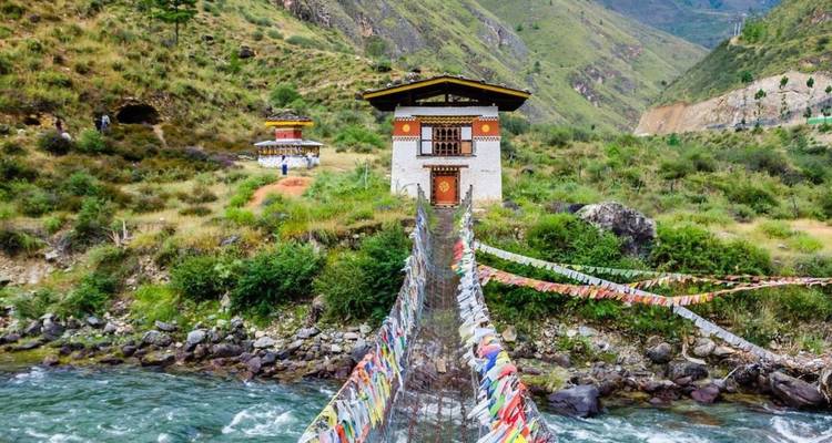 Schmale Hängebrücke mit Gebetsfahnen, die einen reißenden Fluss zu einem kleinen bhutanischen Haus überquert.