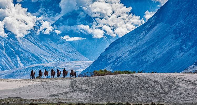 Karawane von Reitern durchquert Sanddüne mit hoch aufragenden blauen Himalaya-Gipfeln im Hintergrund.