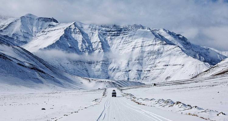 Schneebedeckter Bergpass mit einem einzelnen Fahrzeug, das auf einer weißen Straße auf zerklüftete Gipfel zufährt.