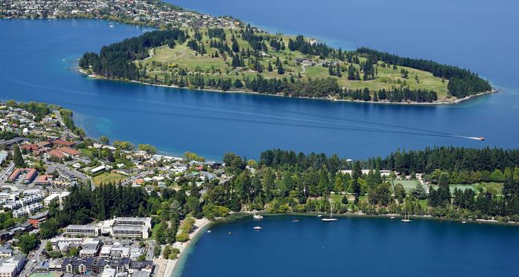 Luftaufnahme einer baumbedeckten Insel und eines tiefblauen Sees, der von Stadtgebäuden und Hügeln umgeben ist.