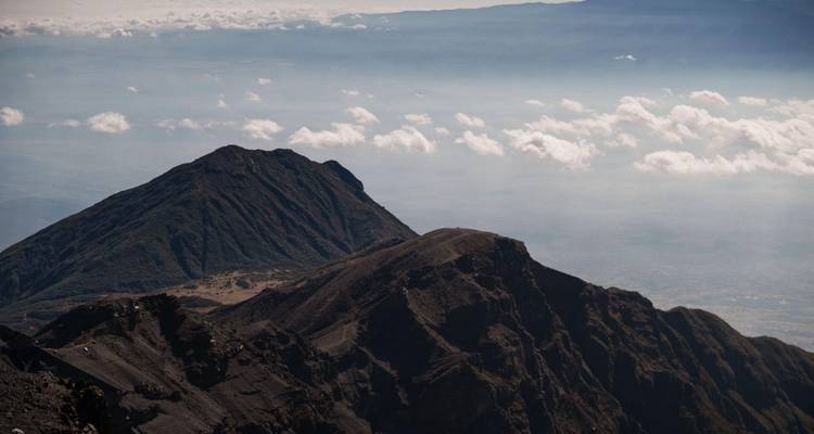 Pics volcaniques sombres et escarpés s'élevant au-dessus d'une mer de nuages avec des plaines lointaines et brumeuses.
