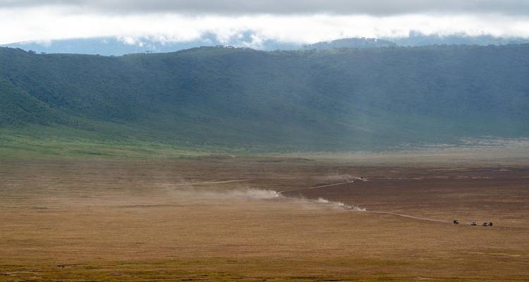 Vue panoramique du fond du cratère du Ngorongoro avec des pistes de safari poussiéreuses et un rebord verdoyant sous de bas nuages.
