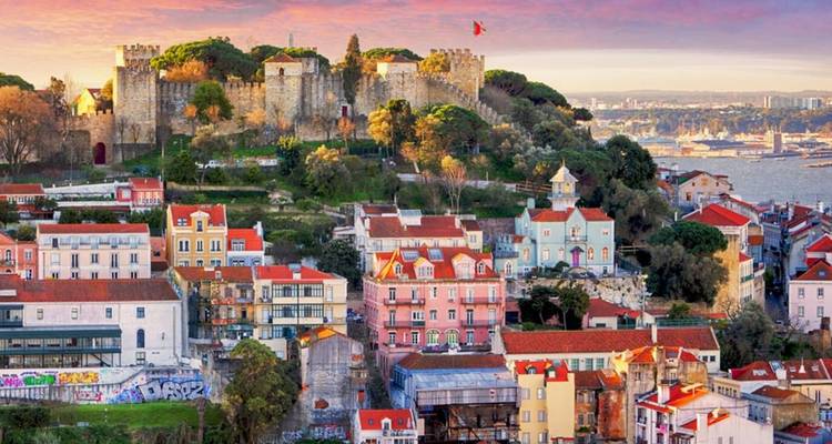 Coloridos tejados de Lisboa ascendiendo hacia el Castillo de São Jorge bajo un cielo de atardecer pastel y la bahía.