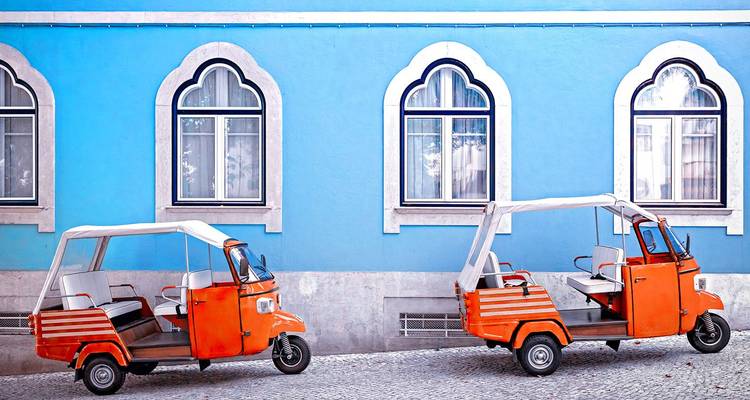 Dos tuk-tuks naranjas brillantes estacionados frente a una pared azul vívida con ventanas arqueadas.