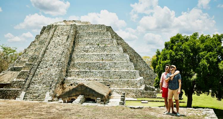 Twee reizigers die poseren voor een oude stenen piramide onder een heldere blauwe hemel.