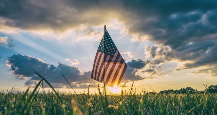 Amerikaanse vlag met achtergrondverlichting door gouden zonsondergangstralen die oprijst boven een grazig veld.