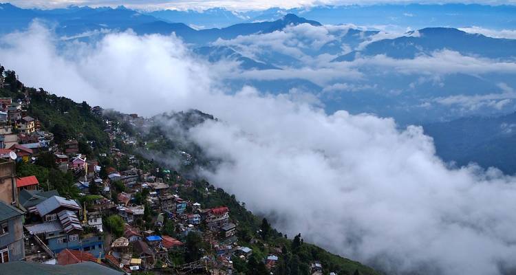 Wolken wälzen sich über terrassierte Hügeldörfer, während entfernte Himalaya-Grate über dem Nebel hervortreten.