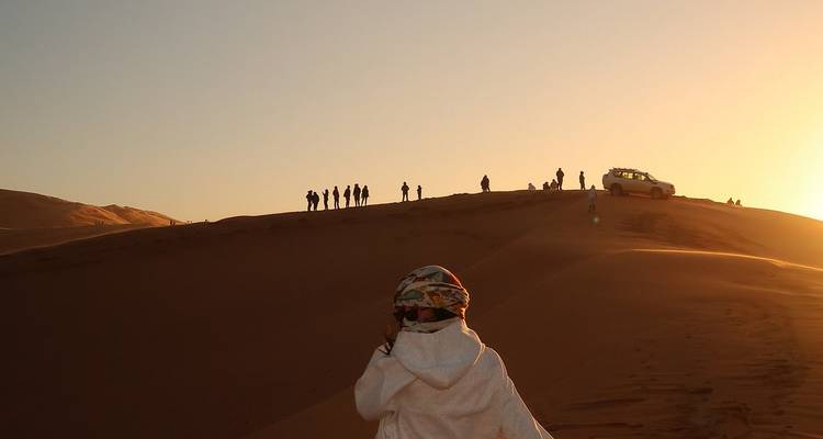 Des silhouettes de voyageurs se dressent au sommet d'une dune de sable éclairée par le soleil tandis qu'une personne en robe les observe et qu'un véhicule 4x4 est garé à proximité au coucher du soleil.