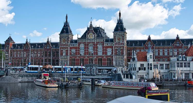 La gare centrale historique d'Amsterdam avec ses tours ornementées se reflétant dans le port aux côtés des tramways et des bateaux de tourisme.