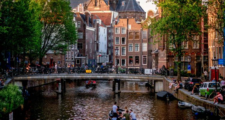 Scène animée des canaux d'Amsterdam avec un pont, des cyclistes, des bateaux et des maisons à pignons étroits dans la lumière chaude du soir.