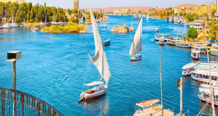 Helderblauwe Nijl met witgezeilde feloekas en levendige waterkant van Aswan.