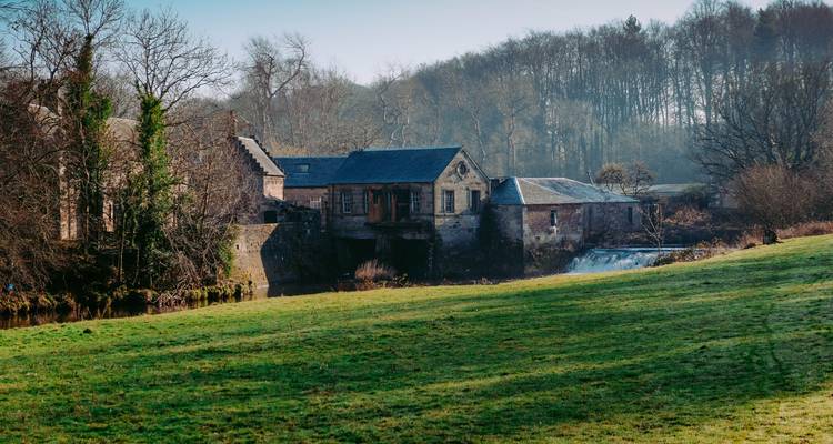 Les bâtiments rustiques d'un moulin en pierre se dressent à côté d'un petit barrage et d'un champ herbeux bordé par des arbres dénudés en hiver dans la brume matinale.