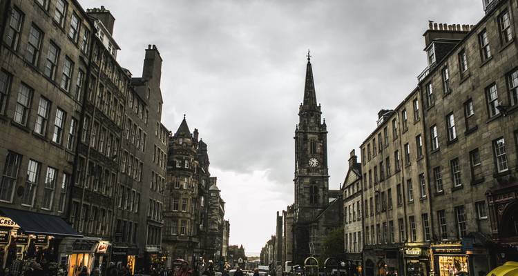 Rue pavée aux teintes sombres de la Vieille Ville d'Édimbourg, bordée de bâtiments en pierre et d'une haute tour d'horloge sous des cieux gris.