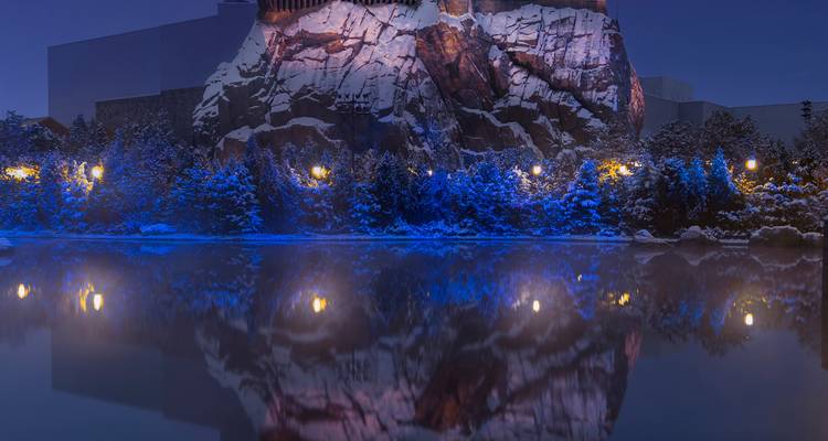 Scène nocturne d'une structure rocheuse dramatique en montagne illuminée par des teintes bleues et violettes se reflétant dans une eau calme.