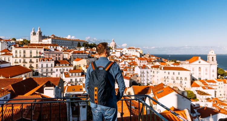 Un joven viajero con mochila contempla el distrito histórico de tejados rojos de Lisboa desde un balcón panorámico con el río Tajo a lo lejos.