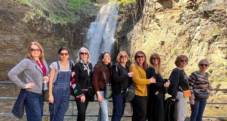 Groep glimlachende vrouwen poserend op een reling voor een rotsachtige waterval.