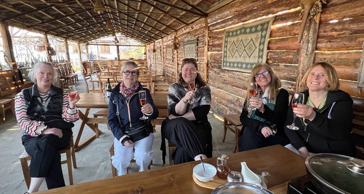 Vijf vrouwen zitten in een houten openluchtlodge, heffen wijnglazen op en glimlachen naar de camera.