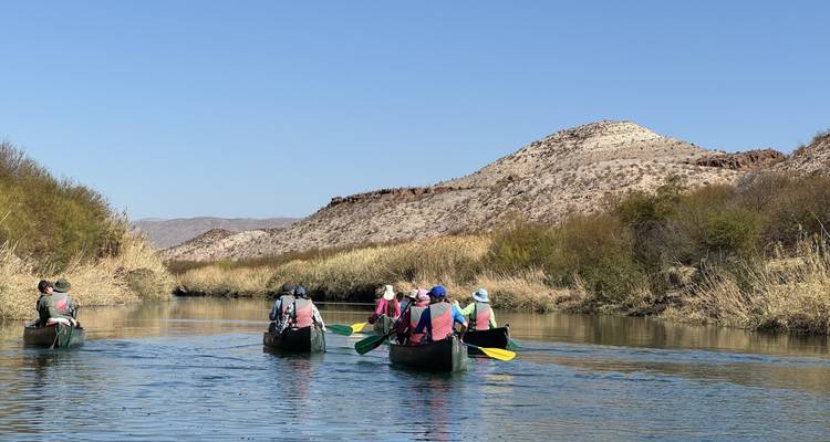 Trois canoës avec des pagayeurs naviguant sur une rivière désertique calme entre des rives broussailleuses