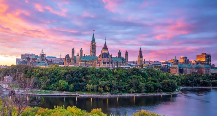 La Colline du Parlement d'Ottawa illuminée par un ciel de lever de soleil coloré au-dessus de la rive bordée d'arbres.