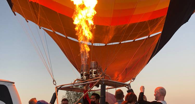 Passagiere stehen in einem Heißluftballon-Korb, während Zwillingsbrennern eine riesige orange Flamme in die farbenfrohe Hülle gegen den Morgenhimmel schießen.