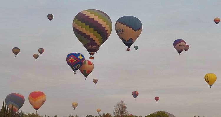 Dutzende bunter Heißluftballons treiben über den grauen Morgenhimmel über einer fernen Landschaft.