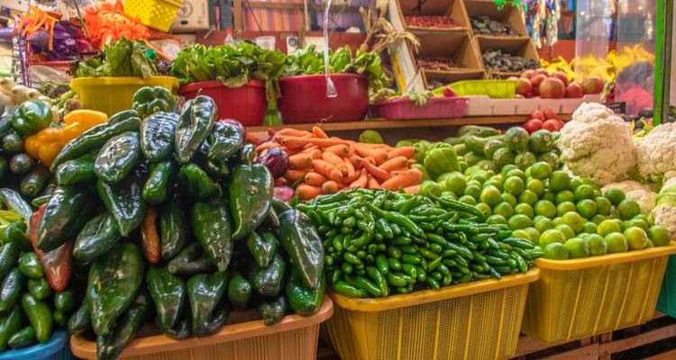 Gehäufte Körbe mit leuchtenden Gemüsesorten und Chilis in der Auslage eines belebten mexikanischen Marktes.
