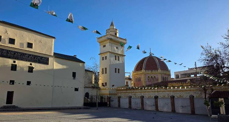 Courtyard with a minaret and domed mosque decorated by strings of Algerian flags under a clear blue sky.