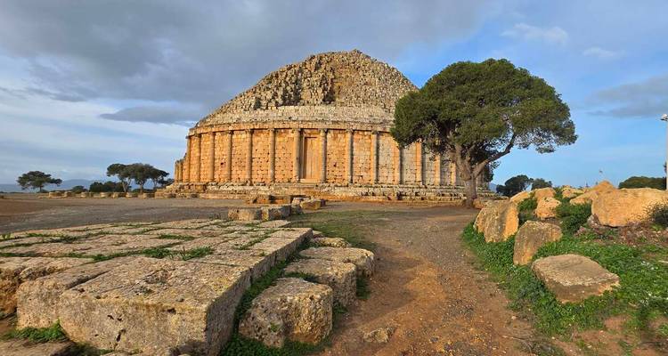 Ancient domed mausoleum built of large stone blocks surrounded by scattered ruins and trees.