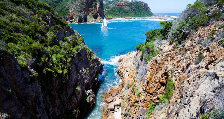 A sailboat glides through turquoise water between steep rocky cliffs at the Knysna Heads inlet.