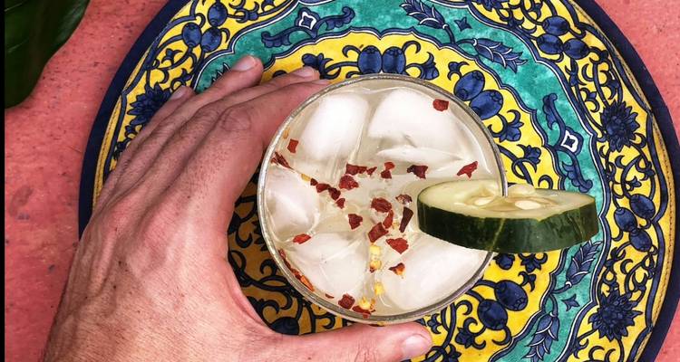 Draufsicht auf einen Cocktail mit Eis, garniert mit Chiliflocken und Gurke auf einem bunten Teller.