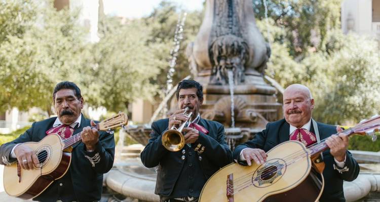 Drei Mariachi-Musiker, die Gitarre und Trompete vor einem Brunnen spielen.