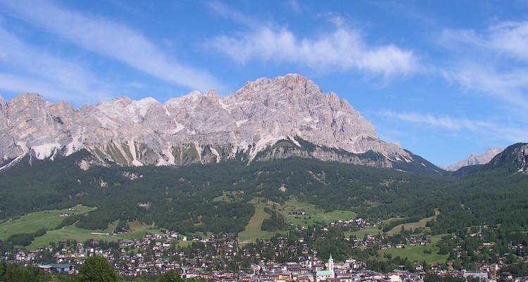 Breed uitzicht op Cortina d'Ampezzo, verscholen onder een rotsachtige Dolomietenrand onder een gestreepte blauwe hemel.
