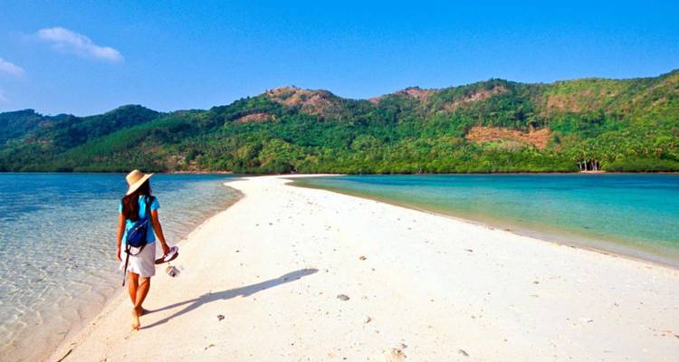 Woman strolls along a pristine white sandbar bordered by crystal-clear blue waters and green hills.