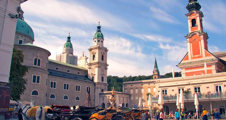 Centre historique de Salzbourg avec les tours de la cathédrale et des calèches sous un ciel bleu.