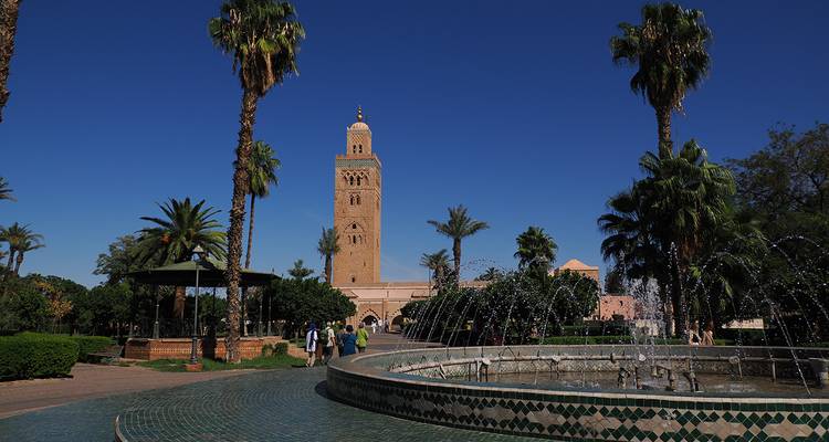La Mezquita Koutoubia se alza detrás de una fuente con azulejos y jardines bordeados de palmeras bajo un cielo azul claro.