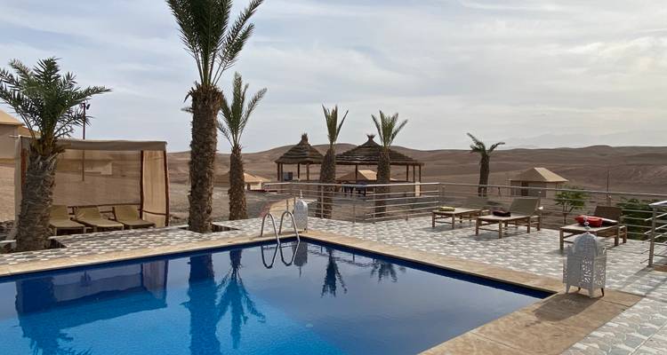 Un campamento boutique en el desierto cuenta con una pequeña piscina, palmeras y pabellones de lona contra colinas áridas.