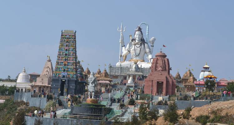 Große Shiva-Statue dominiert Mehrtempel-Komplex mit farbenfrohem Gopuram und Besuchern, die Treppen hinaufsteigen.